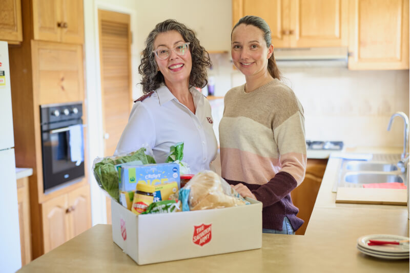 Salvos personnel standing next to a smiling mother with a hamper filled with groceries on the table.