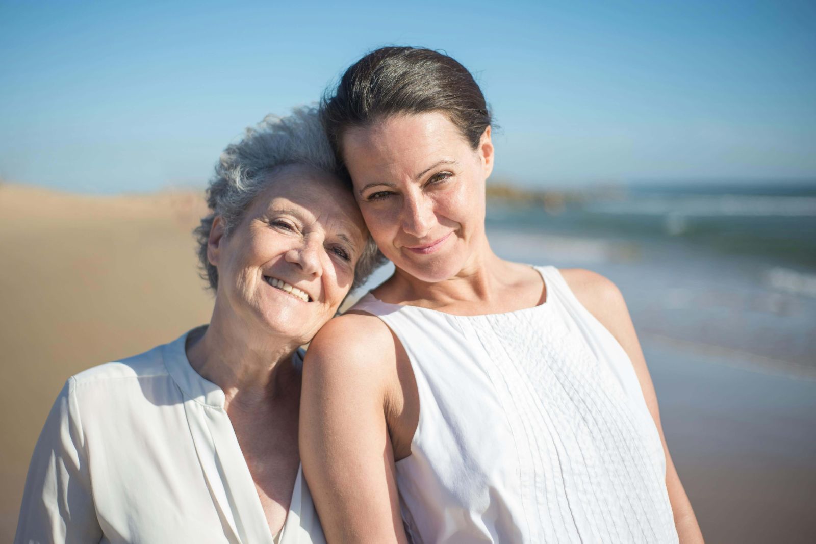 Elder mother and her daughter smiling, standing on the sand in the beach.