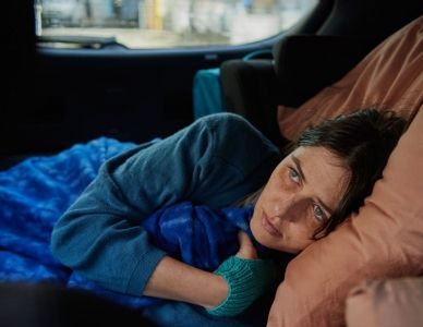 A woman lying on her side in the back seat of a car, wrapped in a blue blanket, looking ahead with a solemn expression.