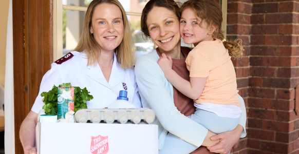 Sophie, a loving mother, is holding her daughter and smiling next to a Salvos personnel holding a Christmas hamper.