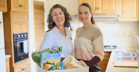 Salvos personnel and Naomi smiling, standing in a kitchen with a hamper filled with groceries on the table.