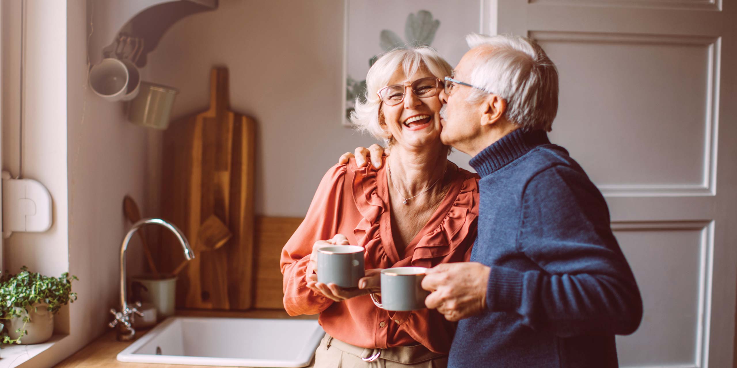 An older couple standing in a cozy kitchen, smiling and enjoying a moment together. The woman, holding a mug, is laughing as the man, also holding a mug, kisses her on the cheek. Wooden cutting boards and plants add warmth to the background.