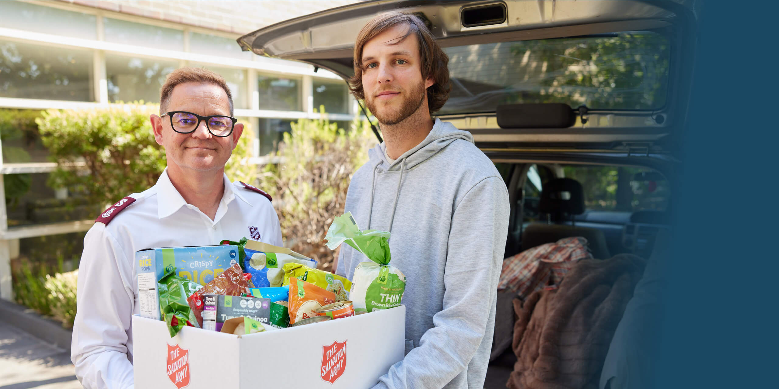 A Salvation Army worker hands a grocery-filled hamper to a smiling man facing financial hardship. Both are smiling, sharing a moment of kindness and support.