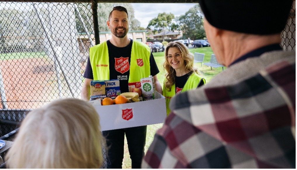 A man and woman from the Salvos stand near a fence, holding a box of food, appearing happy to give to those who need it.